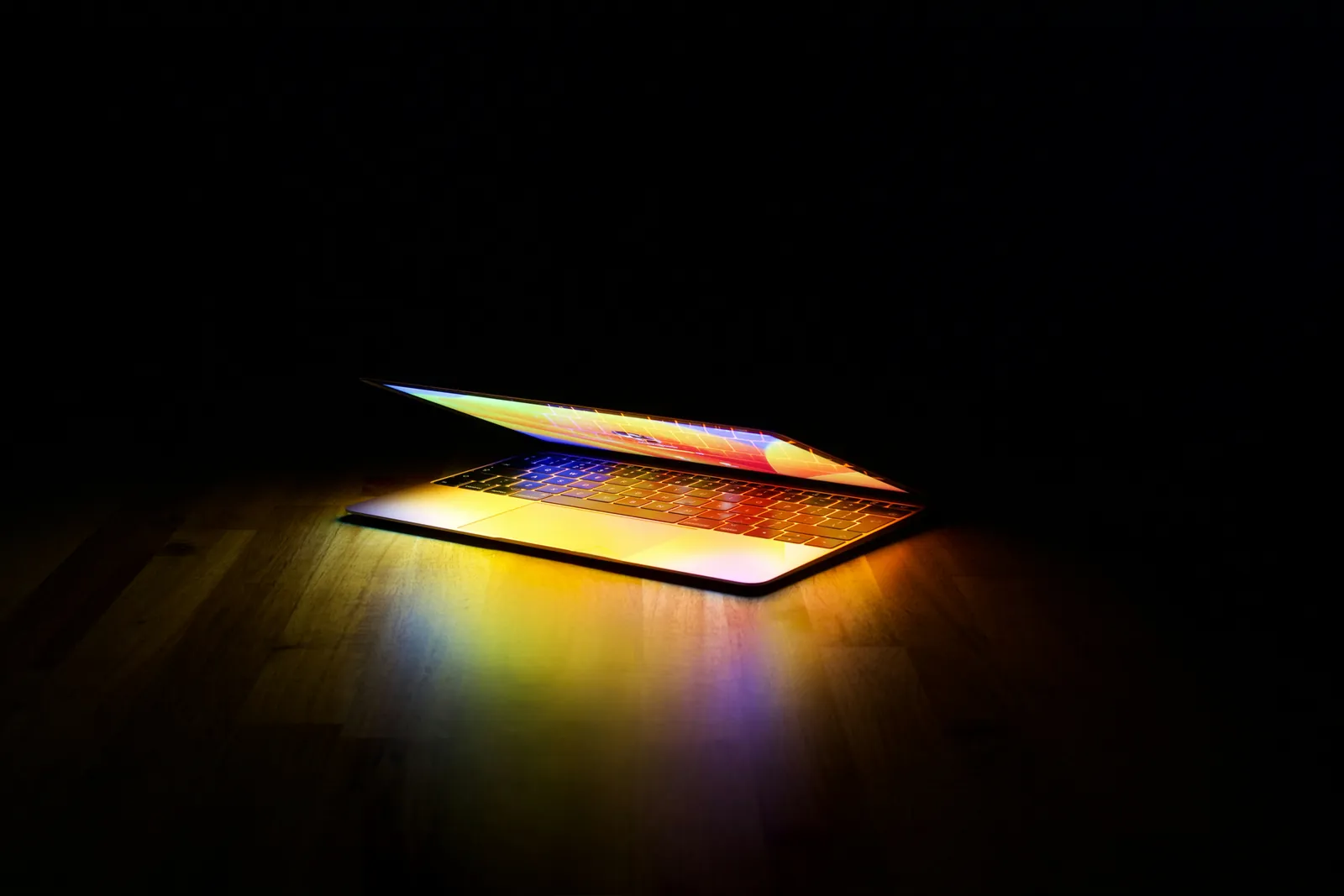 A laptop screen casting colorful light across a wooden desk in a dim room