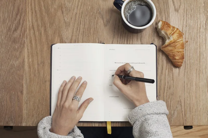 Two hands writing a short list in an open planner beside a cup of coffee