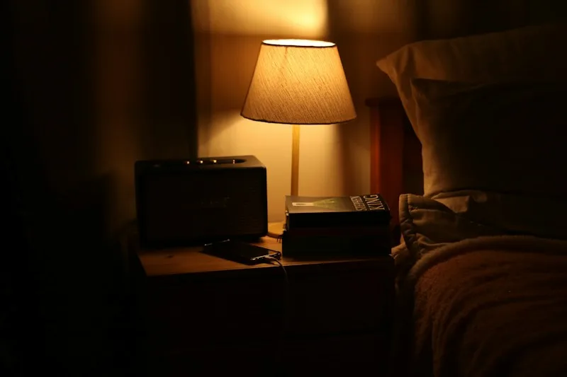 A warm bedside lamp lighting a nightstand in a dark bedroom.