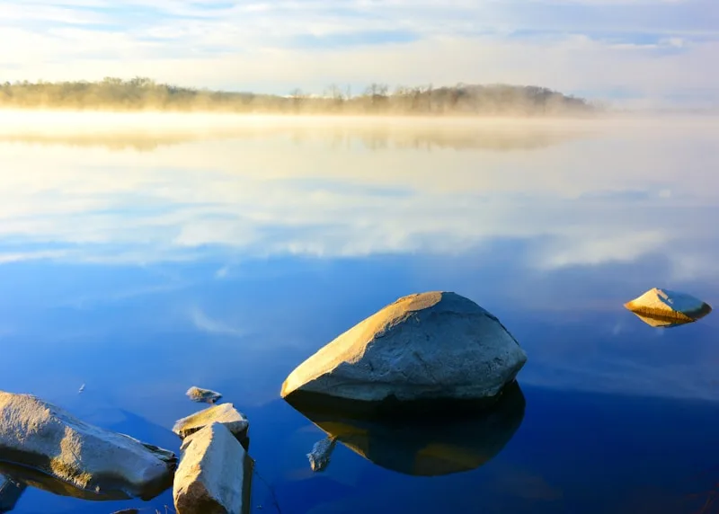 Morning mist lifting off calm, glassy water around a single smooth stone.