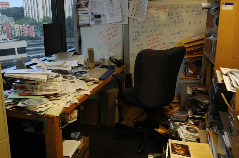 A cluttered office desk piled high with papers and a whiteboard covered in scribbled notes, suggesting scattered, task-switching chaos