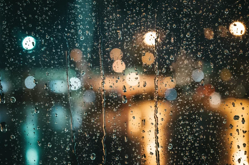 Rain running down a dark window at night with city lights blurred into a soft glow behind the glass.