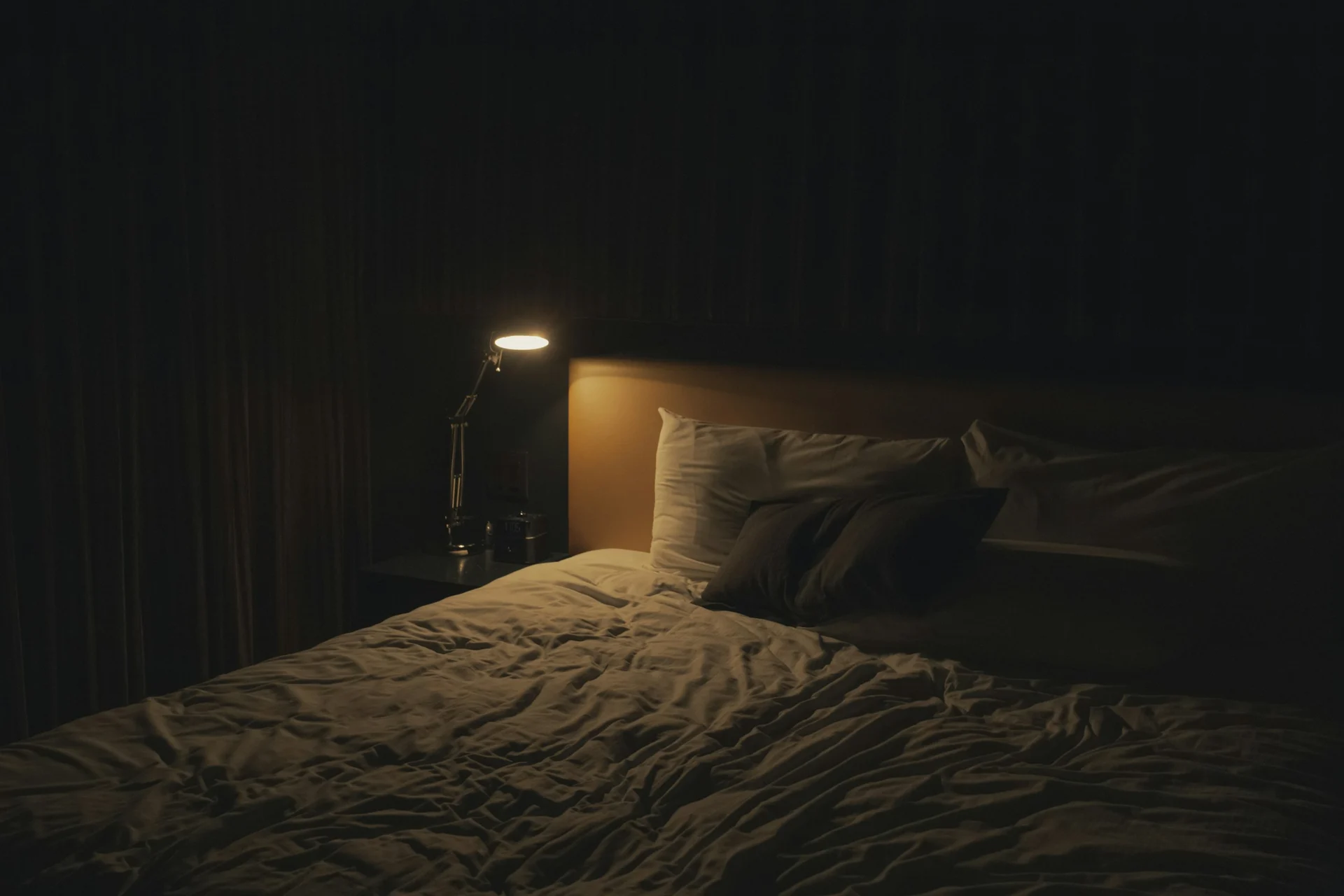 A dark bedroom at night lit only by the soft glow of a bedside lamp over an empty bed