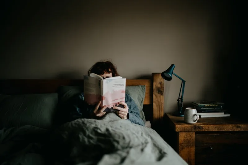 A person reading a paper book in bed by the warm light of a small lamp.