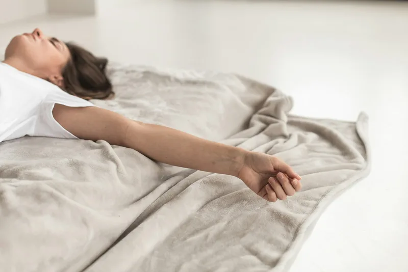 A woman lies on her back resting on a soft grey blanket with her eyes closed.