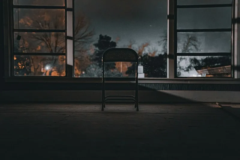 An empty chair facing a window at night, city lights beyond the dark glass