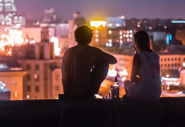 a man and a woman sitting on a ledge overlooking a city at night.