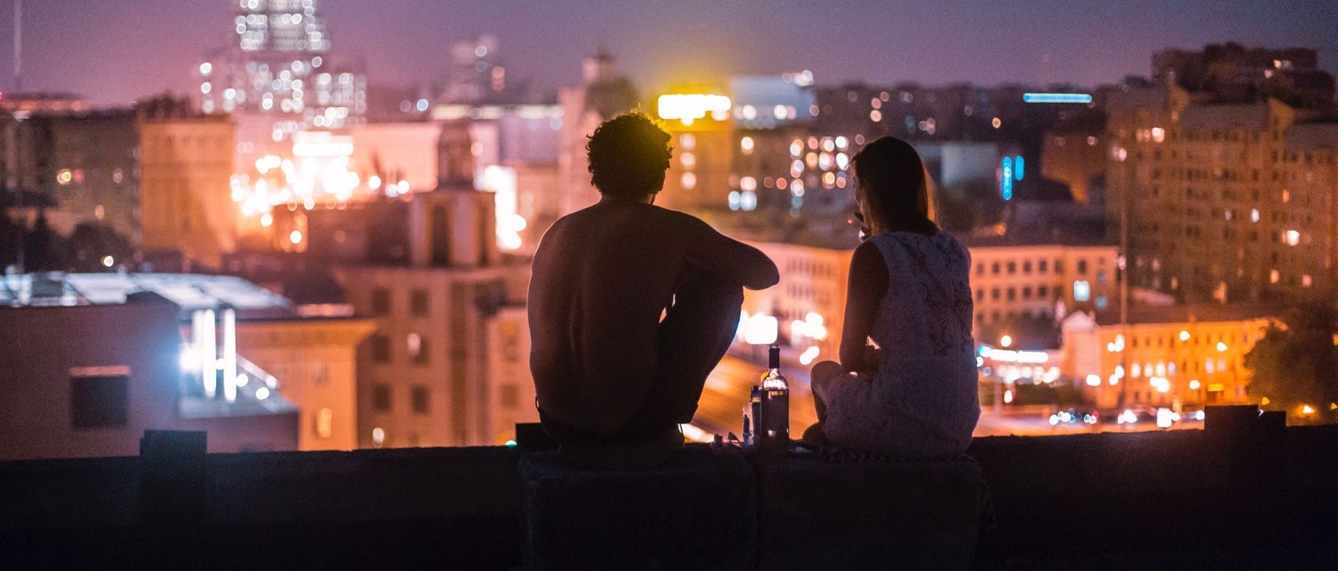 a man and a woman sitting on a ledge overlooking a city at night.