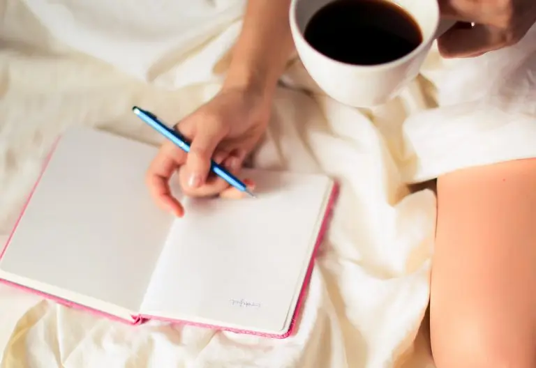 a woman sitting on a bed holding a cup of coffee and writing on a notebook.
