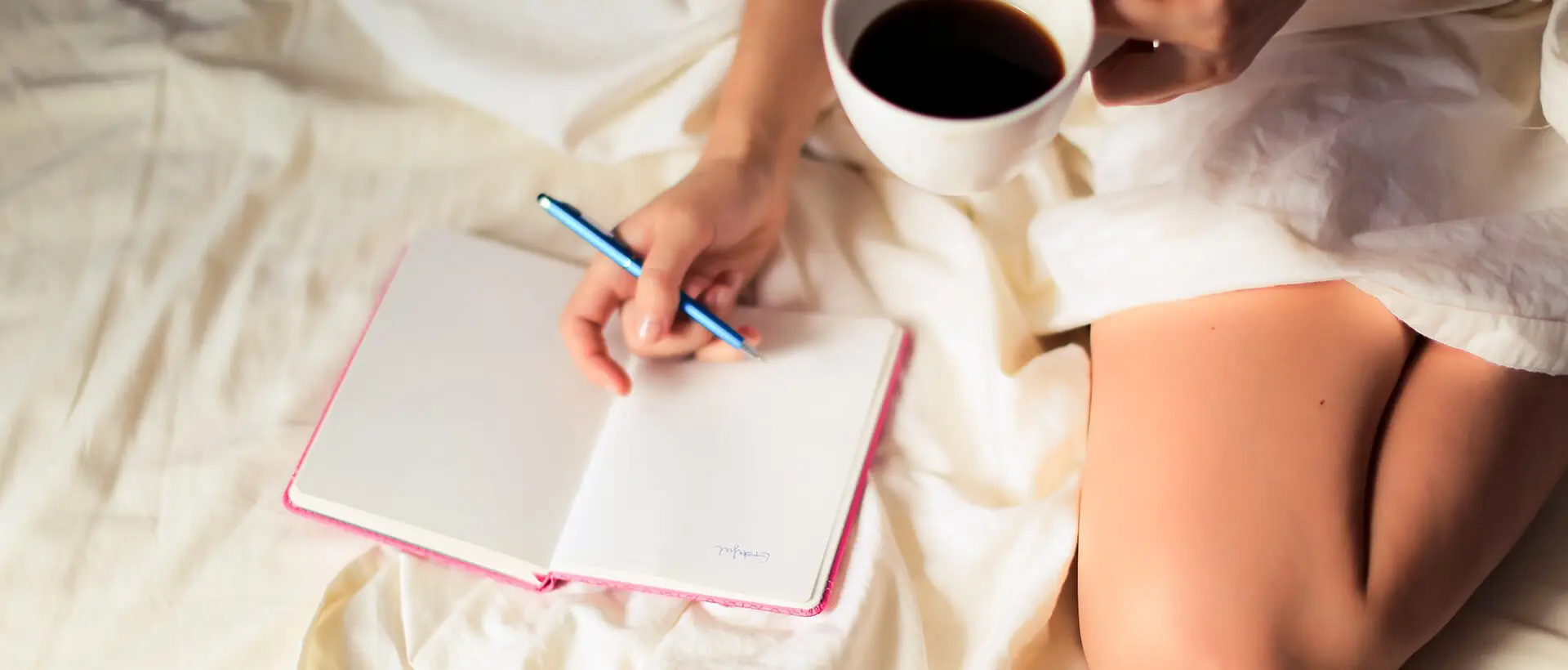a woman sitting on a bed holding a cup of coffee and writing on a notebook.