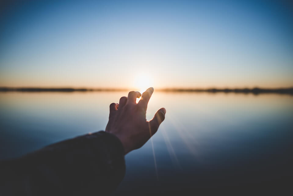 Hand reaching out over a setting sun on water