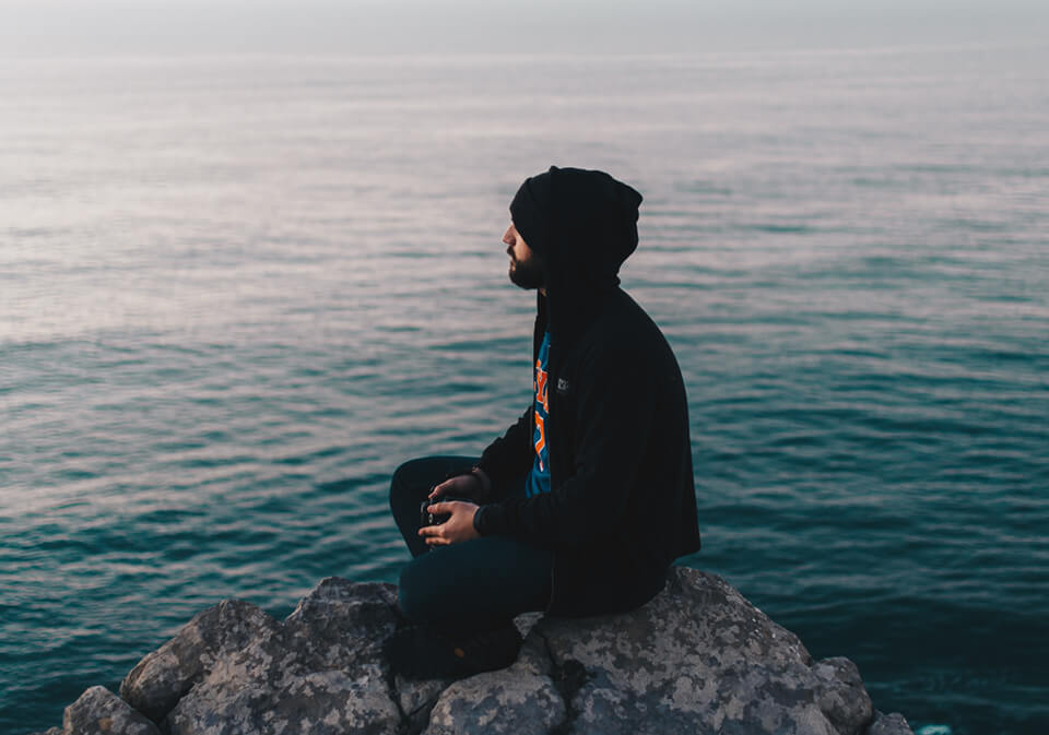 Man with hoodie over head, sitting on rock staring into the horizon